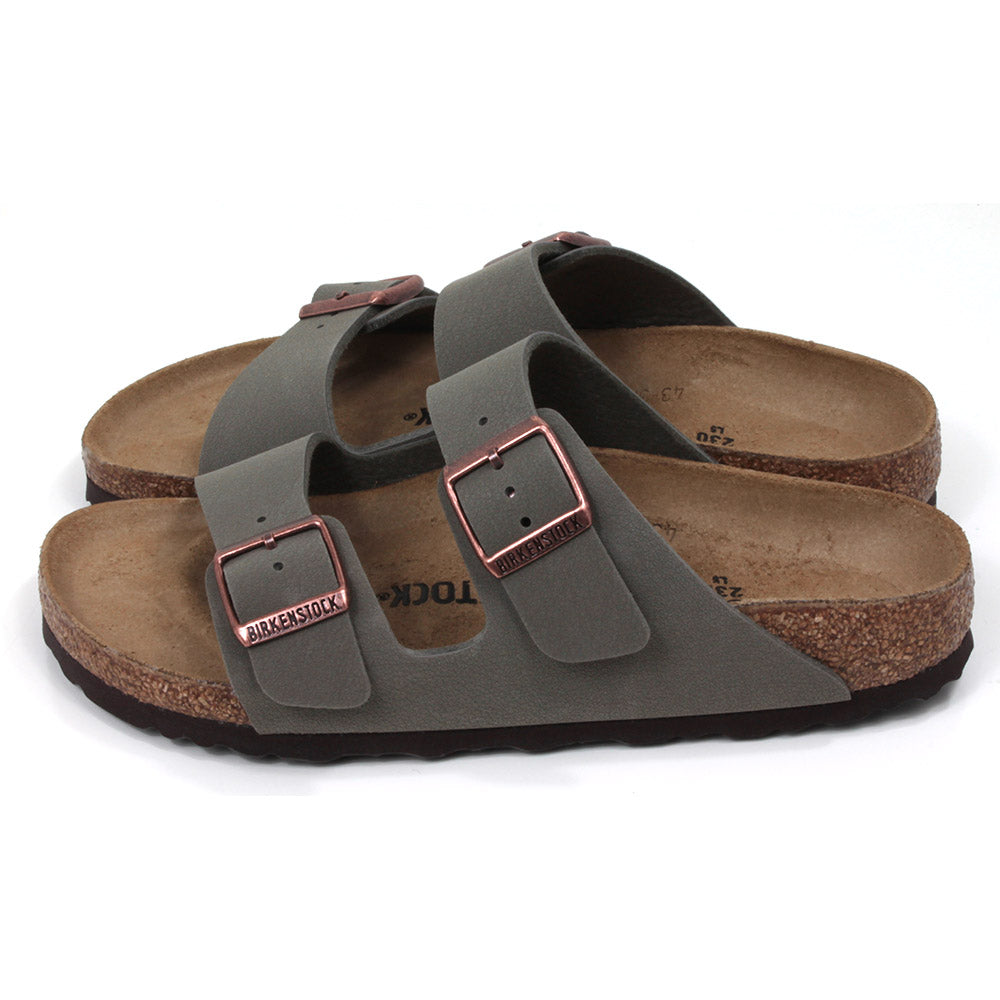 Birkenstock stone coloured Arizona sandals. Two straps over foot adjusted by copper coloured buckles. Open back. Sculpted cork  soles and footbed. Side view.