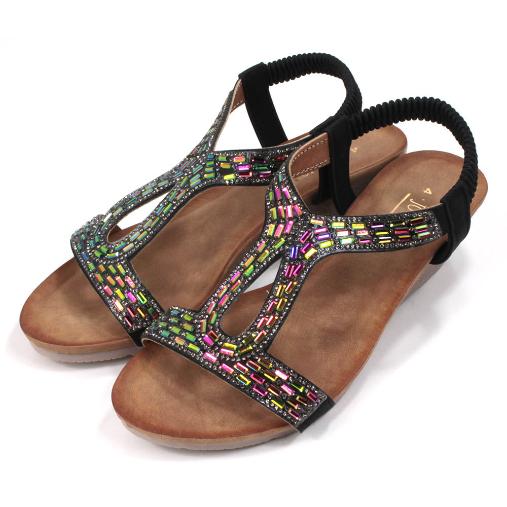 Sandals with black elasticated ankle straps. Over feet straps decorated with mosaics of sparkling jewels. Tan coloured insoles. Low heels. Angled view.