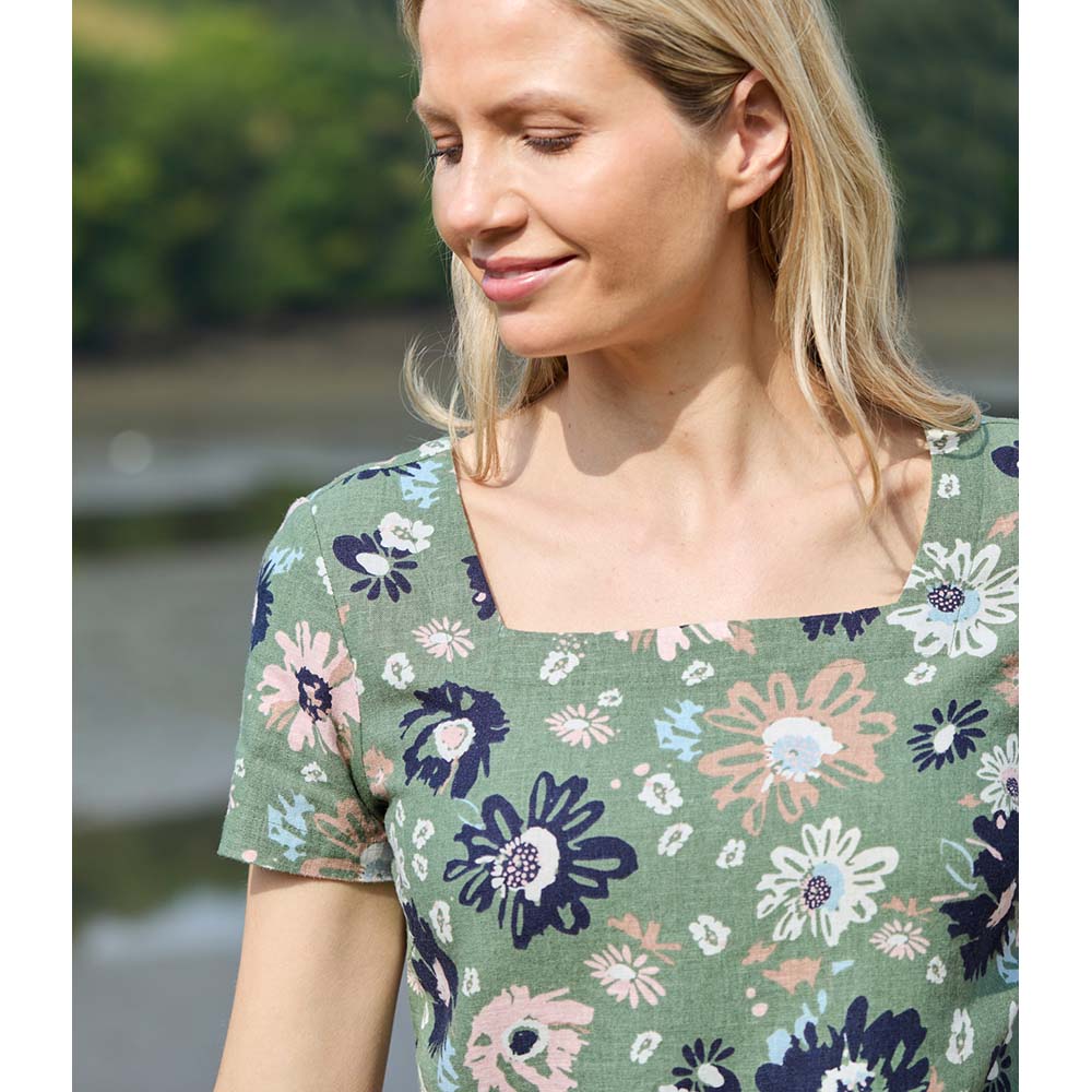 Blonde female wearing a khaki green top that has pale pink and navy blue flowers on it with short sleeves and squared neckline 