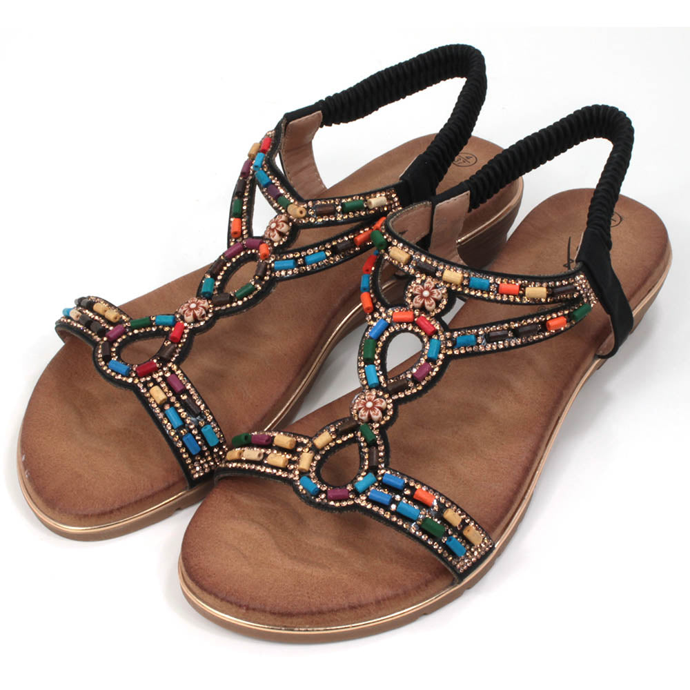 Strappy sandals with elasticated ankle strap in black. Decorated with colourful beads. Tan coloured footbeds. Flats. Angled view.
