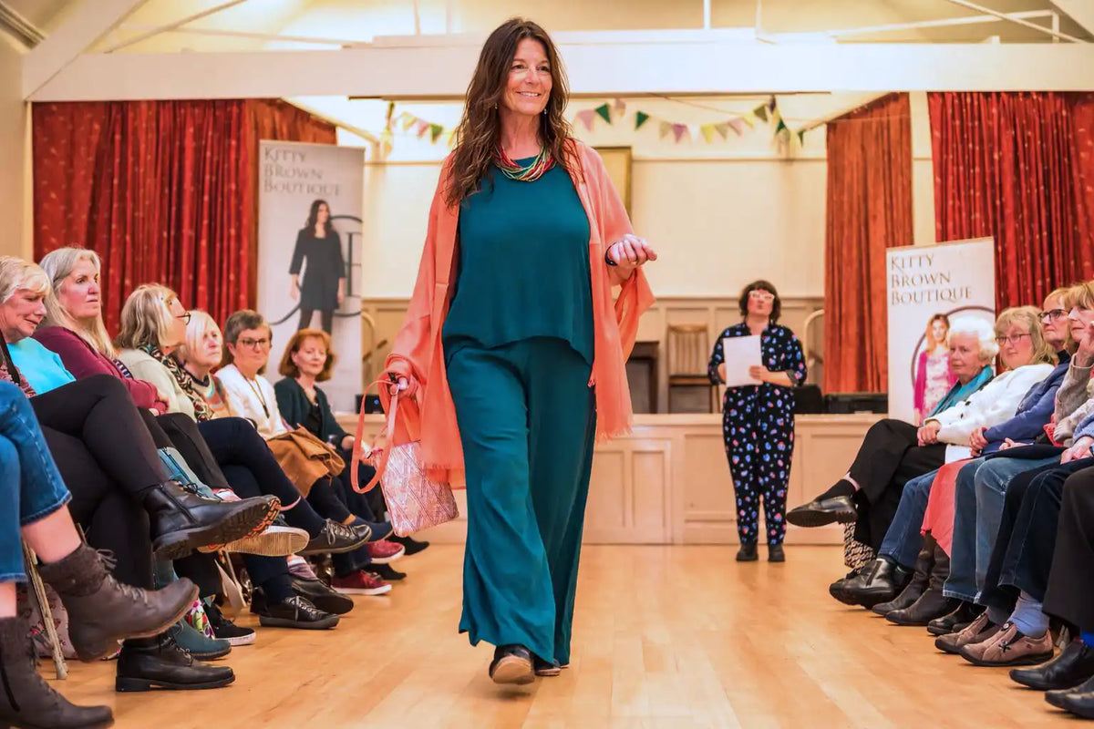 Woman walking down the runway at a Kitty Brown fashion show with the audience looking on.
