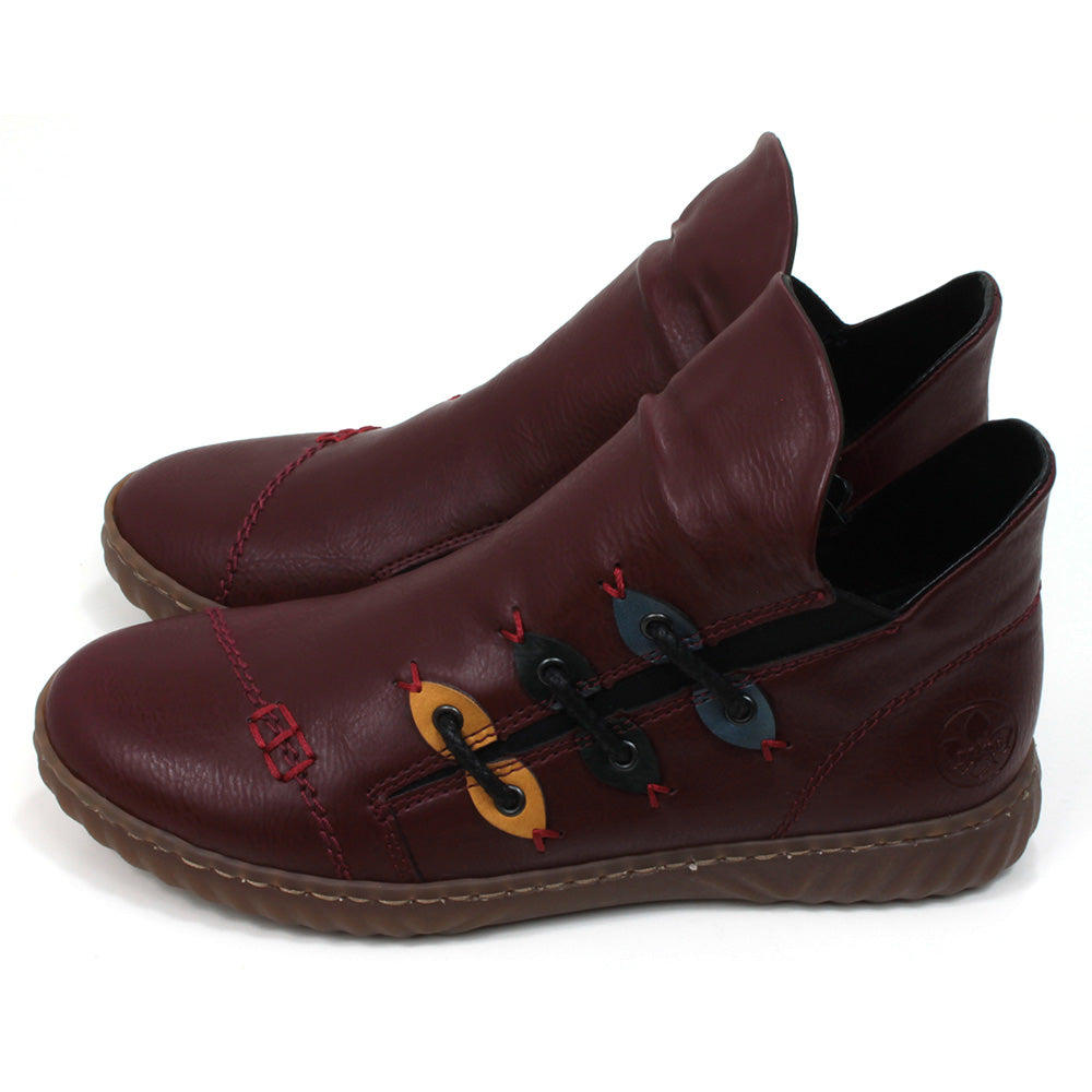 Low ankle boots in wine red colour. Leather effect with red stitching. Zip fitting using black zips. Contrasting colour material details on the sides with faux lacing. Rubber soles. Side view.