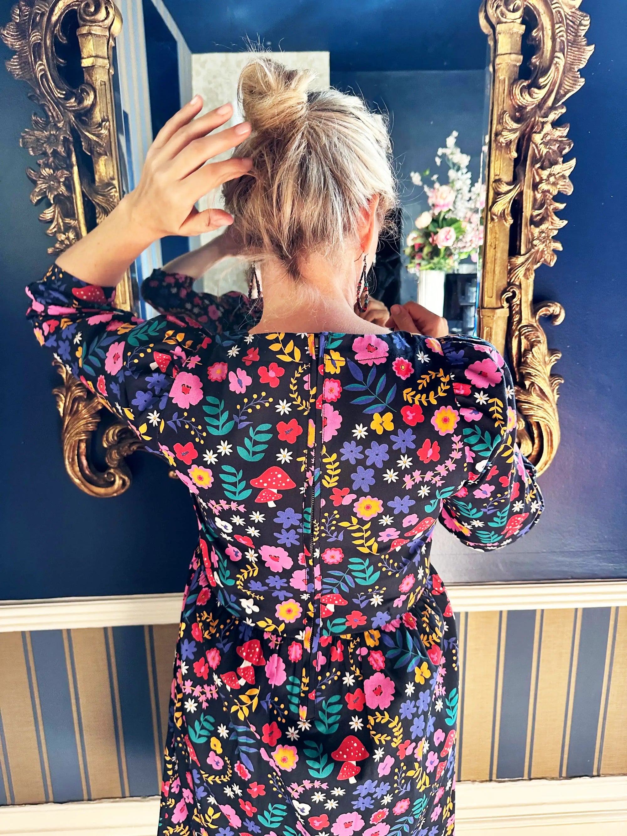 Woman in a floral dress adjusting her hair in front of an ornate mirror.