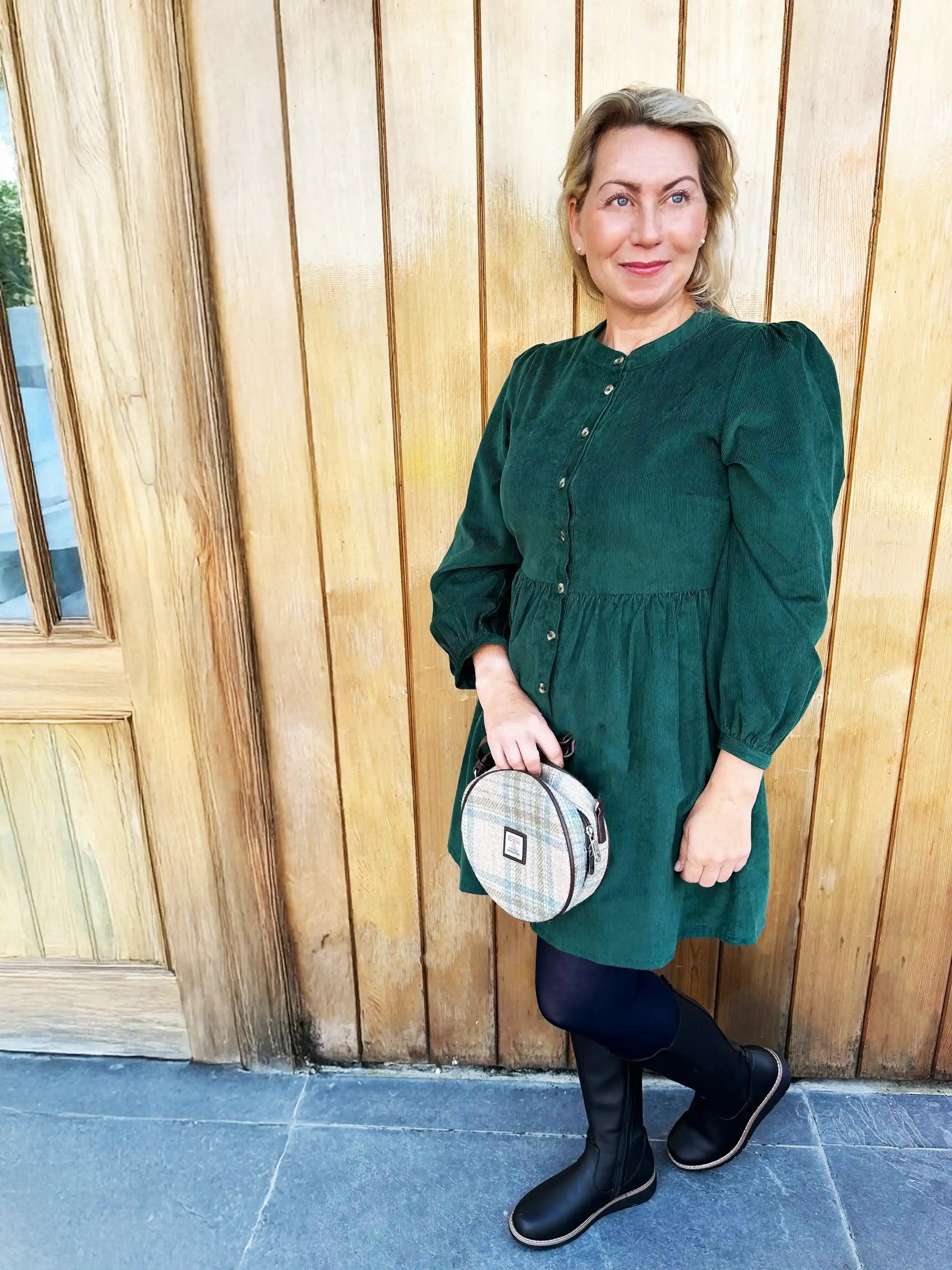 Woman in a green dress holding a patterned handbag against a wooden wall.