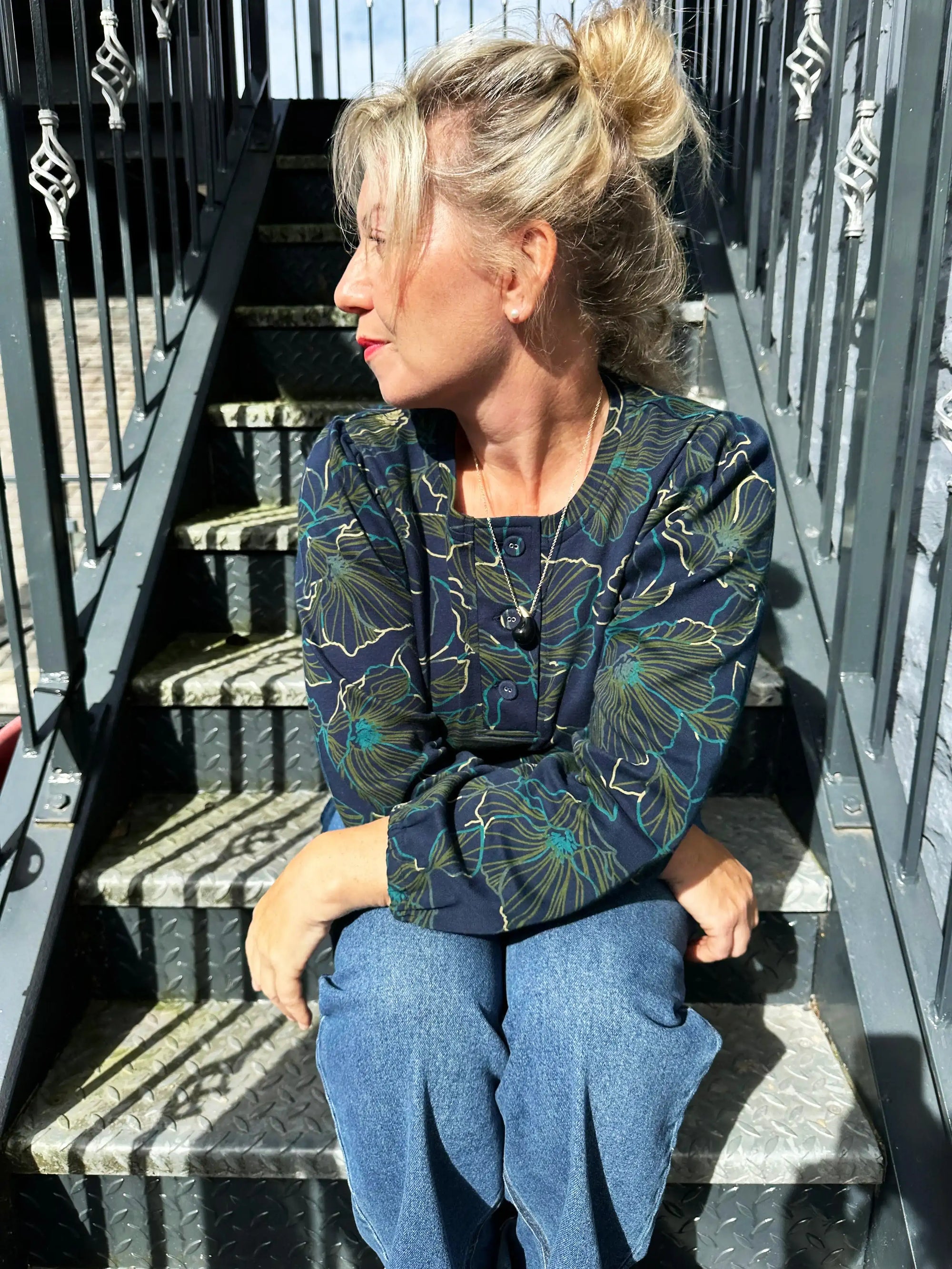 Woman sitting on metal stairs wearing a floral blouse and blue jeans.