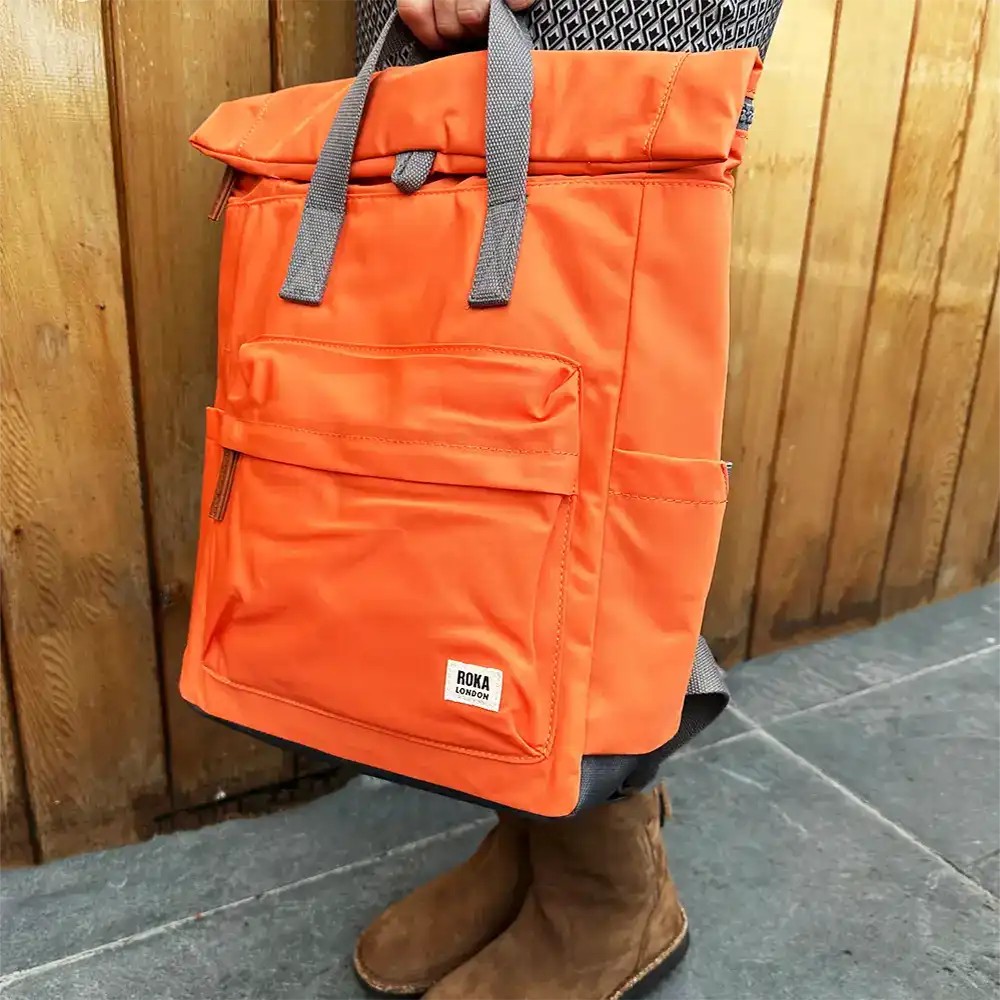 An orange roll-top backpack with gray handles and a small white ROKA label, carried by a person standing in front of a wooden wall.