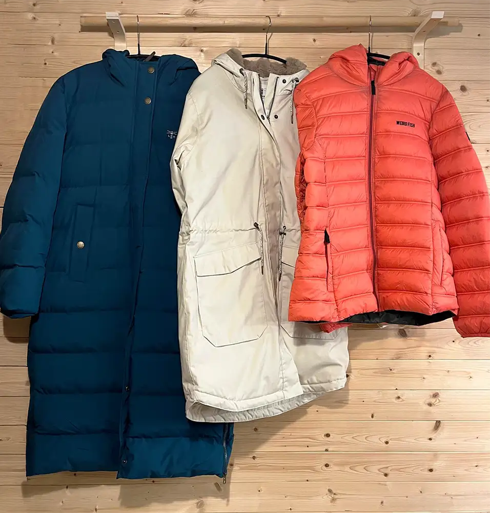 Three puffer jackets in blue, white, and red hanging on a wooden rack.