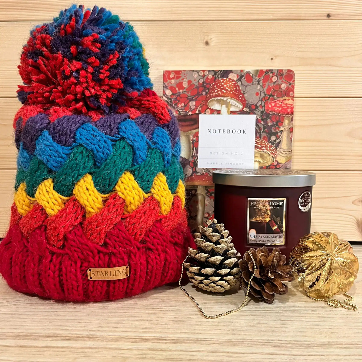 Multi coloured bobble hat, notebook and tinned candle with pine cones and Christmas bauble.