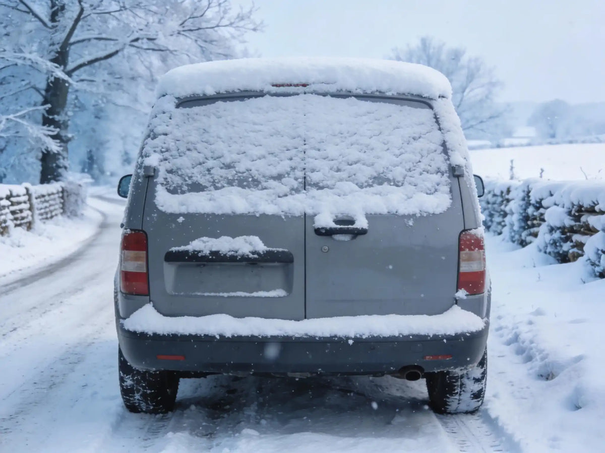 Van covered in snow on a snowy road with trees and a fence in the background