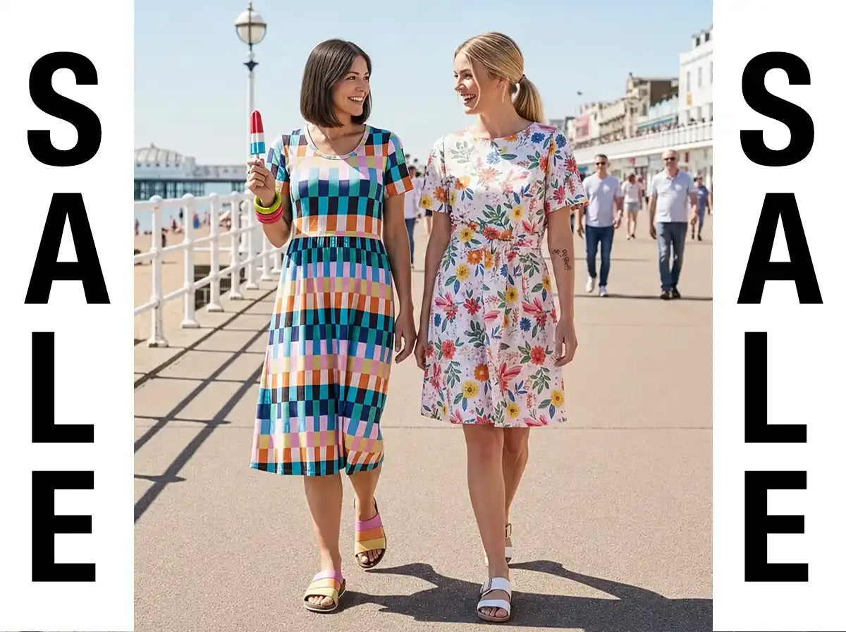 Two women walking hand in hand on a sunny seaside promenade, each wearing a colorful dress. One dress has a blue, orange, and white checkered pattern, and the other has a white base with a vibrant multicolor floral print.