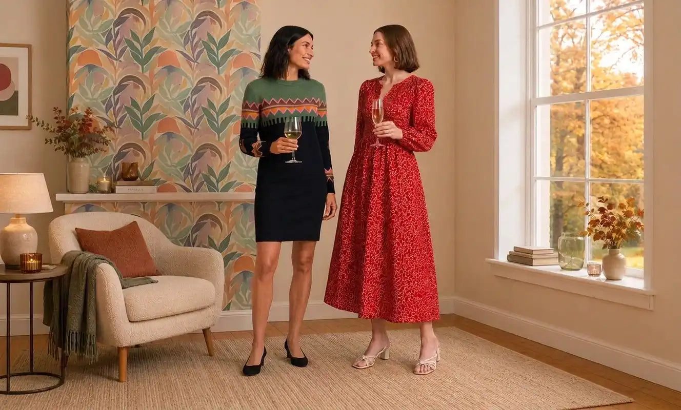 Two women in an elegant room drinking wine. One has dark hair and is wearing a knitted Fair Isle knee length dress. The other has a red A line mid length dress.