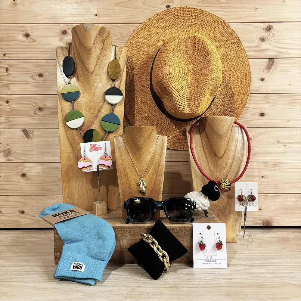 A curated assortment of handmade and streetwear accessories displayed on wooden mannequins and stands against a light wood plank wall. The collection includes a large woven straw hat, a light blue knit beanie with a visible 'Tee Tunes' logo, a pair of black sunglasses, a black velvet pouch with a gold chain bracelet, a pair of red and white beaded earrings, a necklace with circular green, blue, and white beads, and a red headband.