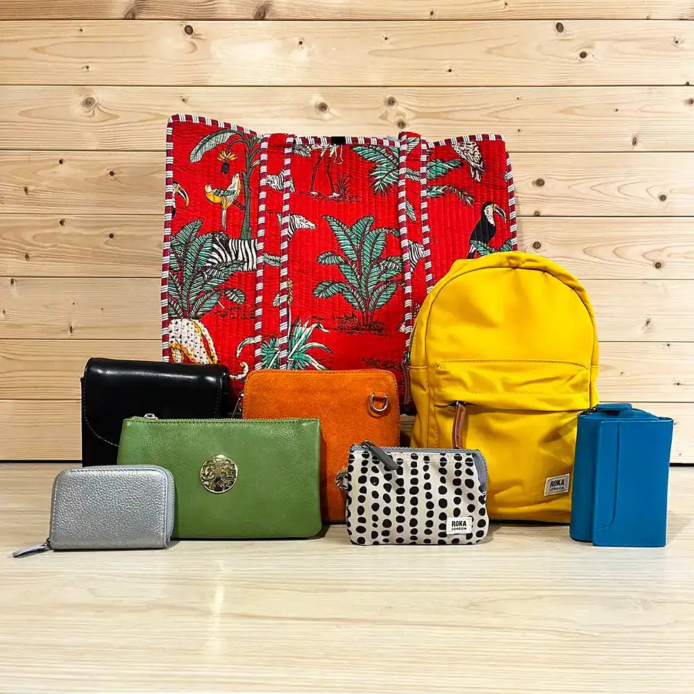 A collection of colorful handbags, wallets, and a backpack arranged in front of a red tropical print tote bag on a light wood plank floor and wall. The items include a yellow backpack, a blue rectangular bag, a black wallet, a green wallet with a gold emblem, an orange pouch, a black-and-white polka dot pouch, and a red patterned tote bag.