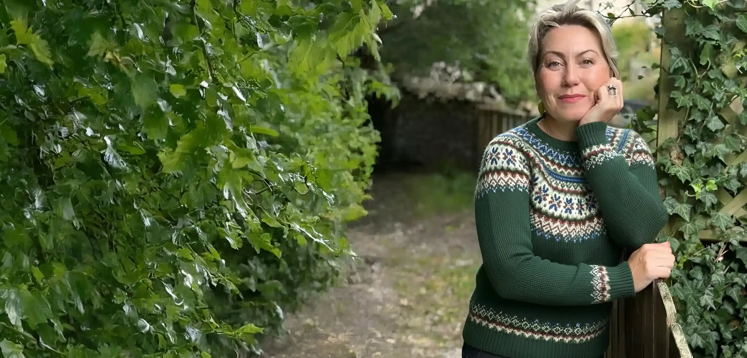 A woman with blonde hair stands on a dirt path surrounded by lush green foliage, wearing a dark green Fair Isle knit sweater with cream and white patterns.