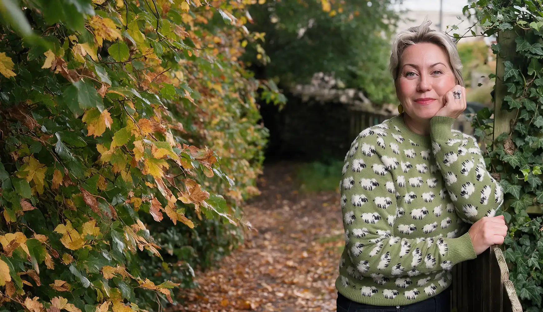 A woman in a green round neck jumper with pattern made of many sheep. She is leaning on a fence. She is beside a leafy hedge with leaves turning yellow for autumn.