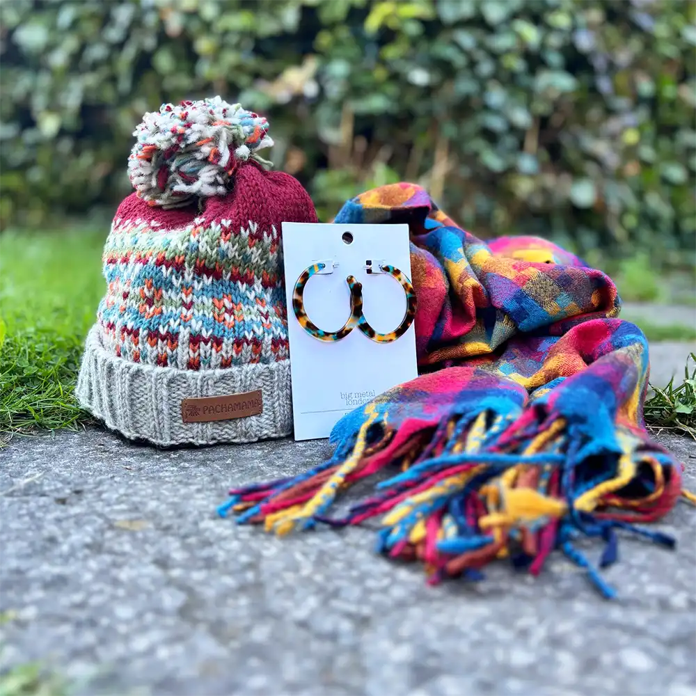 A knit beanie with a multicolor pattern and pom-pom, a colorful plaid scarf, and tortoiseshell hoop earrings on a display card, arranged outdoors on a stone surface with green foliage in the background.