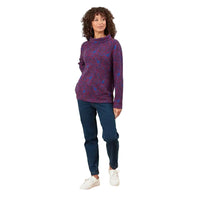 A woman modeling a long-sleeve mock-neck sweater with a red and blue paisley pattern, paired with dark blue jeans and white sneakers, standing against a solid white studio background.