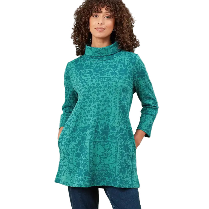 A woman modeling a teal green turtleneck sweater with a tonal floral pattern, paired with dark blue jeans, against a plain white background.