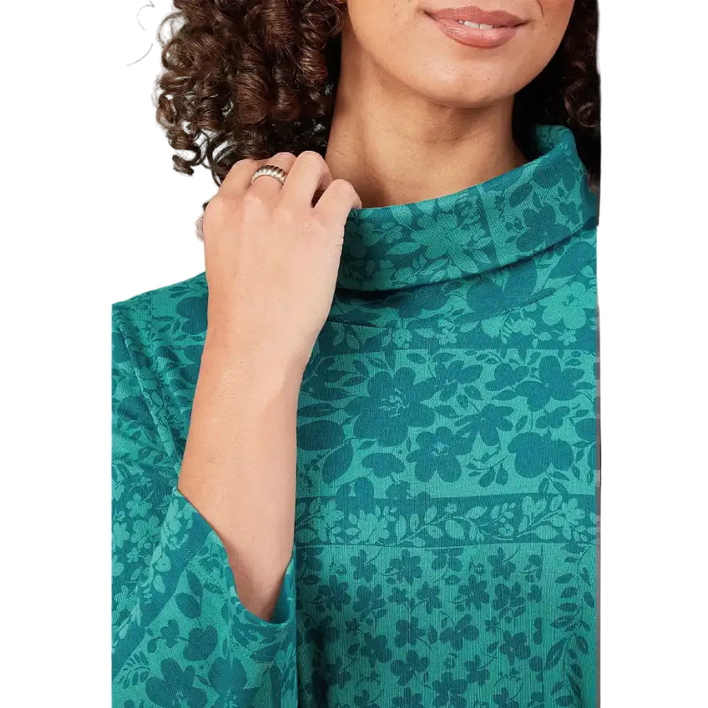Close-up of a woman wearing a teal green floral patterned mock neck top, with her hand resting on her collarbone and a ring on her finger.