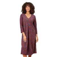 A woman with curly brown hair is shown from the back wearing a long-sleeved midi dress with a dense multicolor abstract print on a muted purple base, set against a white background.