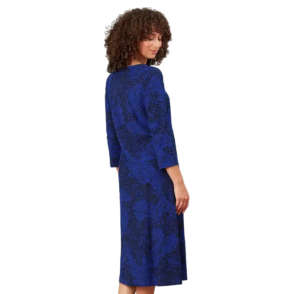A woman with curly brown hair is shown from the back wearing a knee-length blue dress with a dark abstract leaf pattern, set against a solid white background.