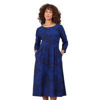 A woman with curly brown hair wearing a long-sleeve blue midi dress with a dark abstract leaf pattern and side pockets, standing against a solid white background.