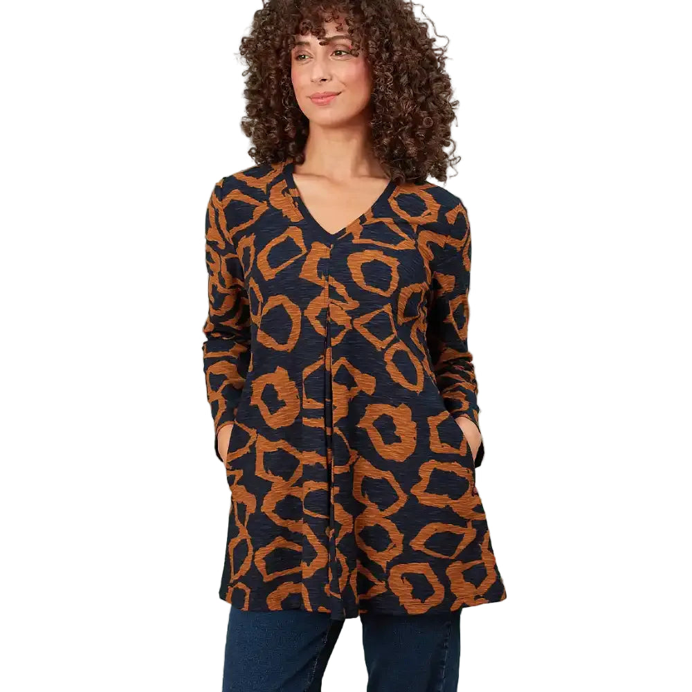 A woman with curly brown hair wearing a long-sleeve V-neck tunic top with a navy blue and rust orange abstract animal print, paired with dark blue jeans, standing in a white environment.