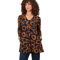 A woman with curly brown hair wearing a long-sleeve V-neck tunic top with a navy blue and rust orange abstract animal print, paired with dark blue jeans, standing in a white environment.