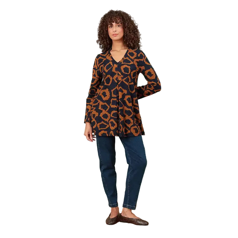 A woman modeling a long-sleeve V-neck tunic top with a navy blue and rust-orange abstract pattern, paired with dark blue jeans and brown shoes, standing in a white environment.