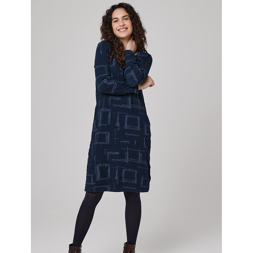 A brunette curly haired model wearing a long sleeve navy blue knee length dress that has a paler blue broken square print on it. with black tights