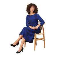 A woman with curly brown hair sits on a wooden chair wearing a blue long-sleeve midi dress with a black abstract pattern, paired with black high heels, against a solid white background.