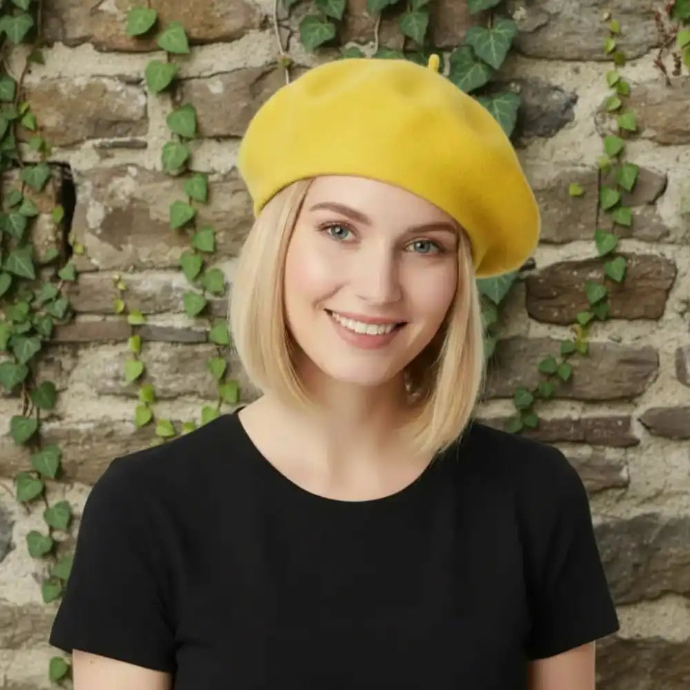 Blond woman wearing a yellow beret and black t shirt in front of a rustic wall with trailing ivy.