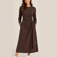 A woman wearing a long-sleeve brown midi dress with a side tie detail and pockets, standing against a plain light beige background.