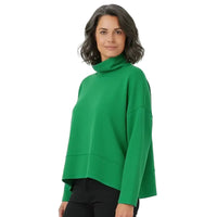 A woman wearing a bright green long-sleeve turtleneck sweater with a relaxed, cropped fit, paired with black pants, against a plain white studio background.