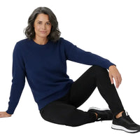 Woman in a relaxed pose on the ground in a navy crew neck jumper and back jeans. She is in a white studio.