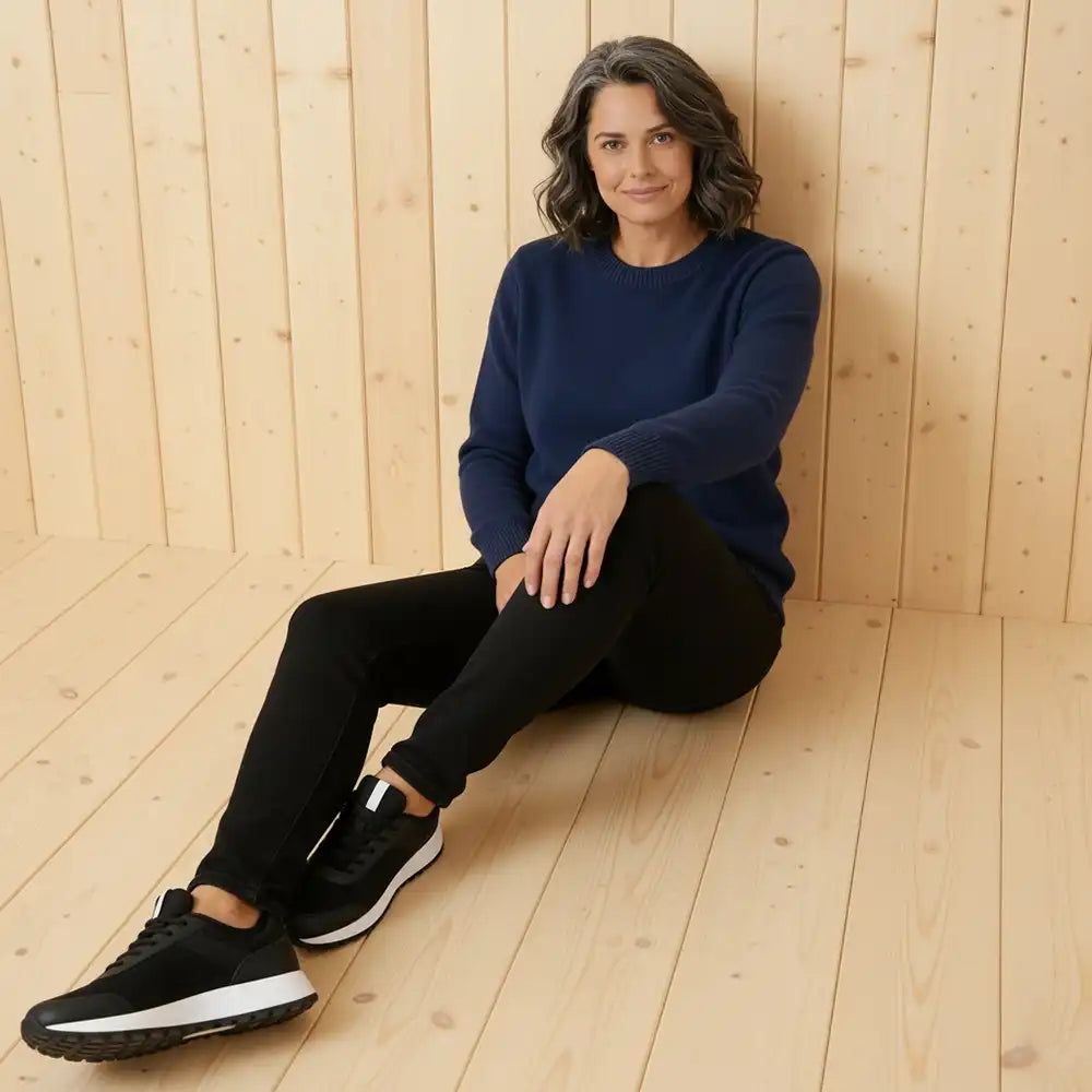 A woman sits on a light wood plank floor against a matching wood-paneled wall, wearing a navy blue long-sleeve top, black pants, and black sneakers with white soles.