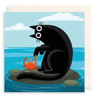 A square greeting card featuring a black cat sitting on a rock in the ocean next to an orange crab, with a cream envelope behind it on a white background.