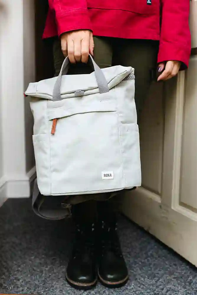 A person wearing a bright red jacket, dark trousers, and black boots holds a light gray canvas roll-top bag with gray handles and a small white ROKA logo patch on the front pocket.