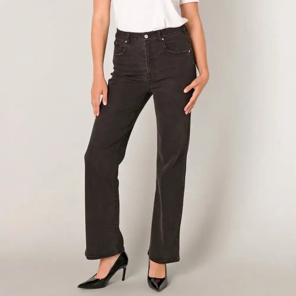 A model wearing a pair of dark charcoal gray wide-leg jeans, styled with a white top and black patent high heels, standing against a light gray studio background.