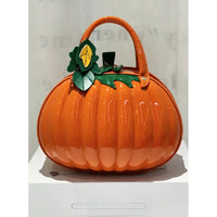 An orange pumpkin-shaped handbag with a glossy finish, featuring green leaf accents and a small yellow leaf detail on the top, displayed on a white pedestal.