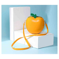 A glossy orange-yellow apple-shaped crossbody bag with a green leafy stem detail, displayed on white geometric blocks against a light blue background.