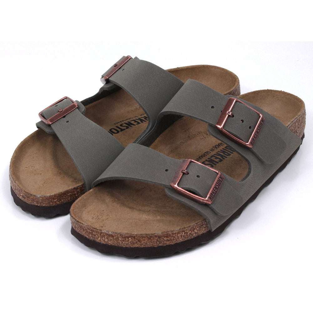Birkenstock stone coloured Arizona sandals. Two straps over foot adjusted by copper coloured buckles. Open back. Sculpted cork  soles and footbed. Angled view.