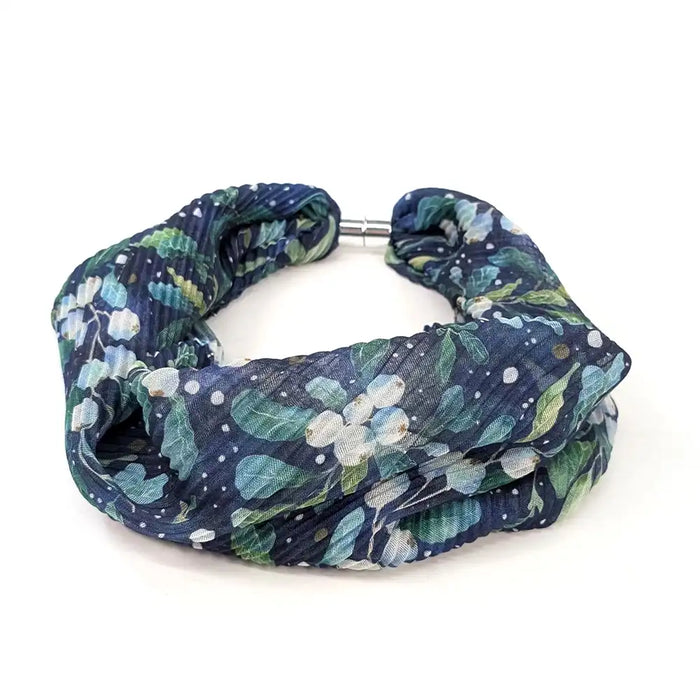 A crinkled fabric headband with a navy blue background, decorated with a green and white floral pattern, and secured with a silver metal clasp, shown on a plain white background.
