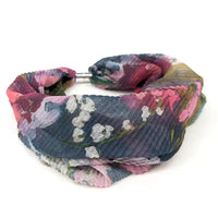 A pleated fabric headband with a colorful floral print, featuring pink, purple, blue, and white flowers on a dark blue-gray background, secured with a silver metal band.