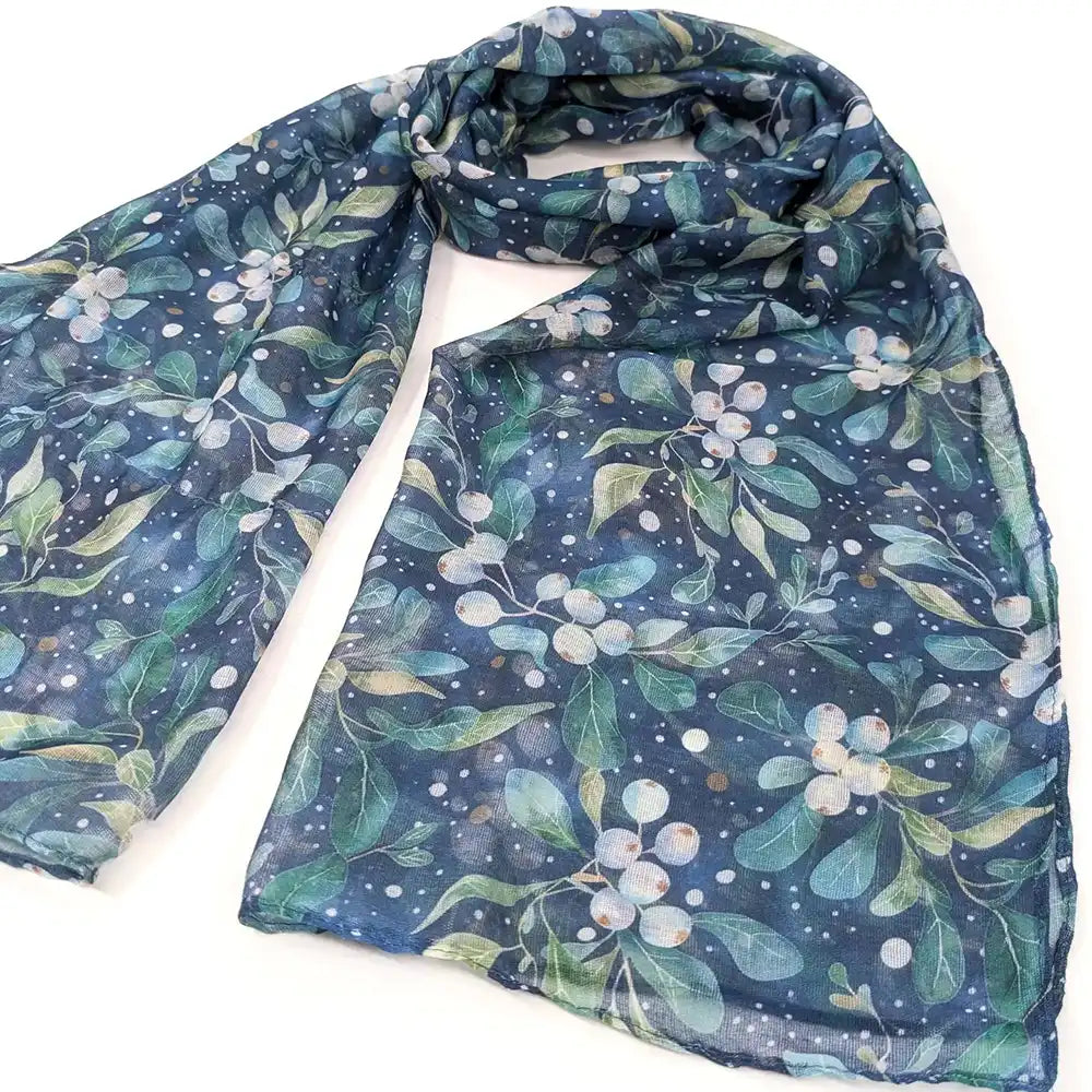 A lightweight scarf with a botanical print of blue-green leaves and white berries on a dark navy background, styled in a loose loop on a white surface.