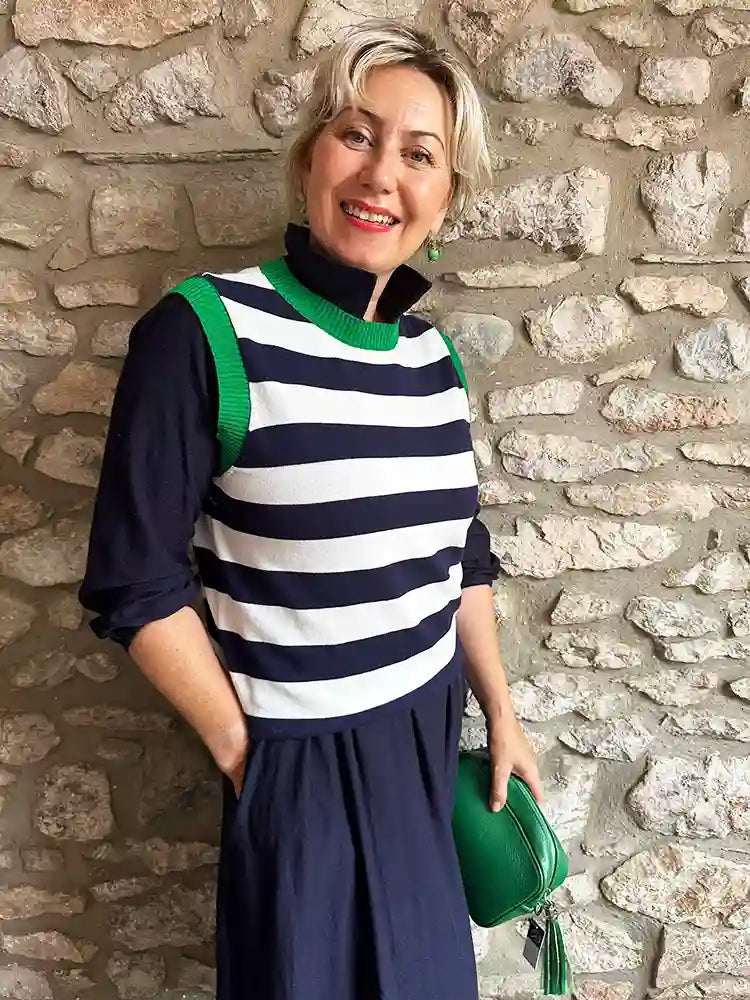 A woman with short blonde hair stands against a rustic stone wall wearing a navy and white horizontally striped mock-neck knit top over a navy long-sleeve dress. She is holding a green handbag.