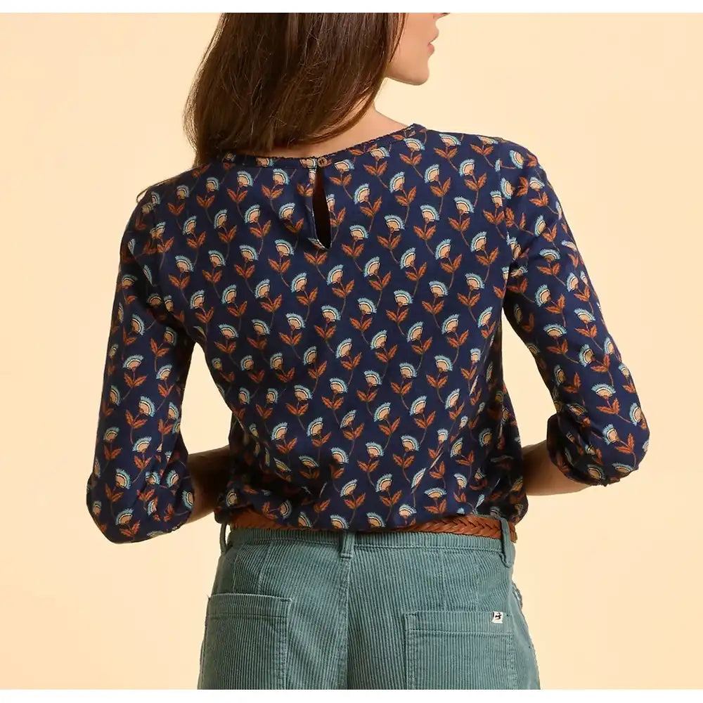 Person wearing a blue patterned blouse with green pants on a beige background