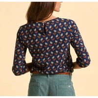 Person wearing a blue patterned blouse with green pants on a beige background