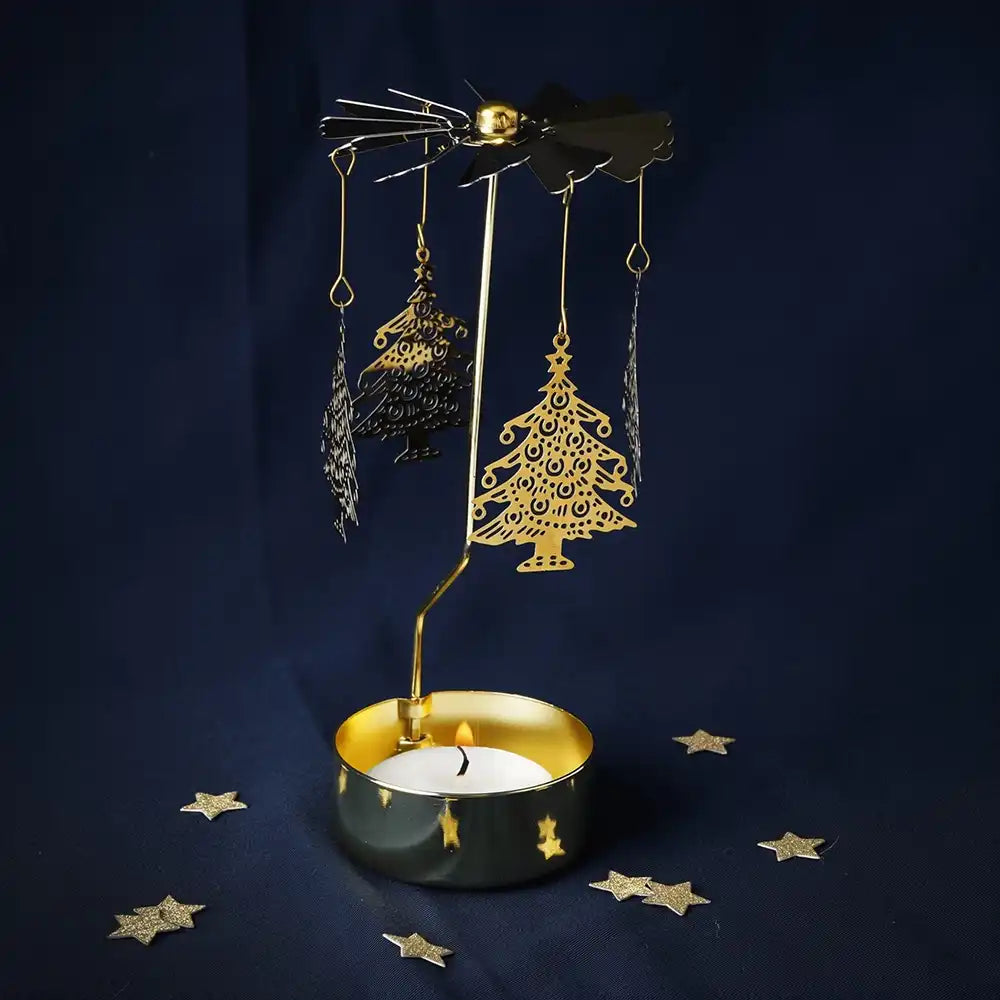 A holiday-themed candle holder featuring a gold metal stand with hanging Christmas tree and bird cutouts, a black candle tin with gold accents, and a lit white candle, set against a dark navy blue background with scattered gold star decorations.