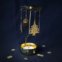 A holiday-themed candle holder featuring a gold metal stand with hanging Christmas tree and bird cutouts, a black candle tin with gold accents, and a lit white candle, set against a dark navy blue background with scattered gold star decorations.